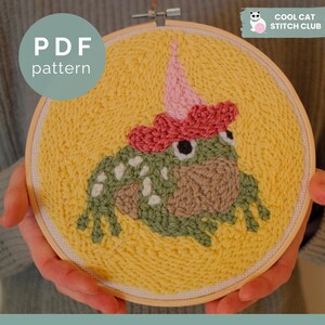 May include: A punch needle embroidery project in a circular wooden hoop featuring a green frog with white spots wearing a pink party hat on a yellow background. The text PDF pattern and Cool Cat Stitch Club are visible on the image.