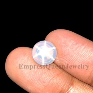 May include: A round, pale gemstone with a six-pointed star pattern. The gem is held between fingers against a black background. The text "EmpressQueenJewelry" is visible below the gem.