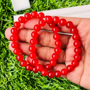 Puede incluir: Dos pulseras de cuentas rojas. Cada pulsera está hecha de cuentas rojas redondas y brillantes. Las pulseras se muestran en una mano sobre un fondo de hierba verde.