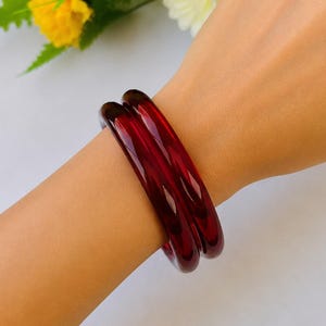 Pode incluir: Duas pulseiras vermelho escuro são usadas no pulso. As pulseiras são lisas e brilhantes, refletindo a luz. O fundo inclui flores amarelas e brancas e folhas verdes.