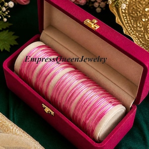 May include: A collection of pink bangles presented in an open magenta velvet jewellery box. The bangles are arranged on a cream-coloured cushion. The text "EmpressQueenJewelry" is printed on the cushion.