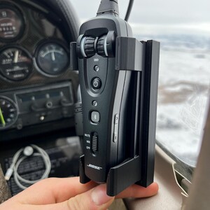 May include: A black Bose aviation headset mounted in an aircraft cockpit. The headset is secured in a black holder. The headset has a power button, Bluetooth indicator, and volume controls. The background shows gauges and a window with a cloudy sky.