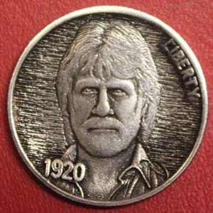 May include: A silver-colored coin featuring a detailed portrait of a man with long hair and a mustache. The word "LIBERTY" is inscribed along the top edge, and the year "1920" is at the bottom. The coin has a textured background.