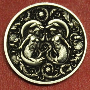 May include: A round, silver-toned button with an intricate design. Two mermaids face each other, surrounded by dolphins and ornate scrollwork. The button has a detailed, raised relief pattern and a dark background.