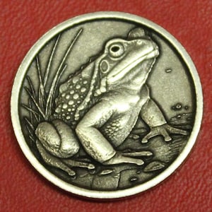 May include: A detailed silver-colored coin featuring a detailed relief of a frog. The frog is depicted with textured skin, sitting near tall grass. The coin's edge is raised, and the background is subtly detailed.