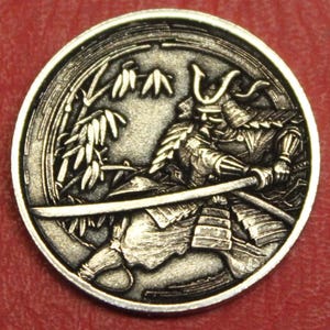 May include: A silver-colored coin featuring a detailed relief of a samurai warrior in full armor, holding a katana. The background includes bamboo stalks. The coin has a circular shape and a detailed border.