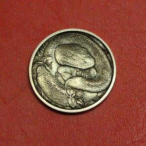 May include: A silver-colored round coin featuring a detailed relief of a coiled snake. The snake's head is prominent, with intricate scales and a focused gaze. The coin's design includes floral elements, all set against a red background.