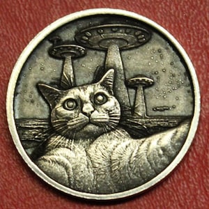 May include: A silver-colored coin featuring a cat with large eyes, seemingly taking a selfie, with three UFOs in the background. The coin has a detailed, engraved design, with a circular shape and a raised edge. The cat's fur is textured, and the UFOs have a classic flying saucer shape.