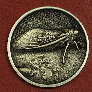 May include: A circular silver-toned metal coin featuring a detailed relief of a cicada insect resting on a textured surface. The insect has prominent wings with visible vein patterns and legs, set against a background of fine etched lines.