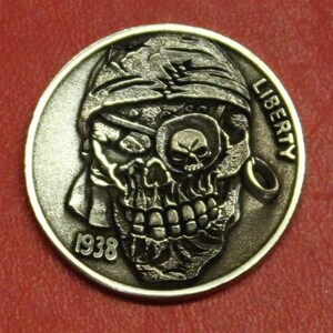 May include: A silver-colored coin with a pirate skull design. The skull has an eye patch and a bandana. The word "LIBERTY" is visible, along with the year "1938". The coin is on a red surface.