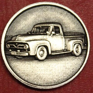 May include: A silver-toned coin featuring a detailed relief of a classic pickup truck. The truck is depicted in profile, showcasing its front grille, headlights, and bed. The coin has a circular shape with a raised border, giving it a vintage feel.