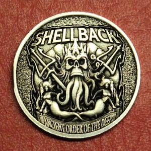 May include: A silver-colored round coin with the word "SHELLBACK" at the top. The coin features a skull with tentacles, tridents, and mermaids. The text "ANCIENT ORDER OF THE DEEP" is at the bottom.
