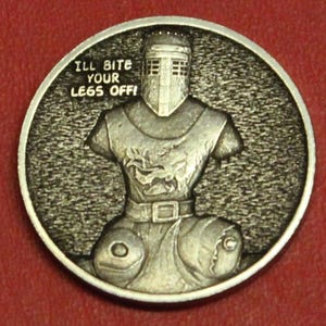May include: A silver-colored round coin featuring a medieval knight with the text "I'LL BITE YOUR LEGS OFF!". The knight is depicted from the chest down, with detailed armor and a helmet.