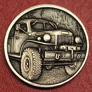 May include: A detailed silver coin featuring a vintage off-road truck. The truck has large tires, a prominent front grille, and a winch. The background shows a forest scene. The coin has a circular shape and a detailed, embossed design.