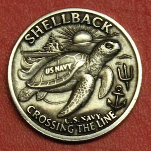 May include: A silver-colored coin with the words "SHELLBACK" and "CROSSING THE LINE" encircling a detailed design. The coin features a sea turtle, a ship, a sun, an anchor, and a trident. The words "US NAVY" are also present.
