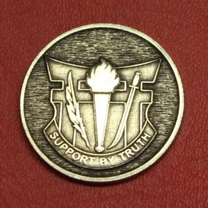 May include: A silver-colored round emblem with a torch, feather, and sword design. The words "SUPPORT BY TRUTH" are inscribed around the emblem's edge. The emblem is set against a red background.