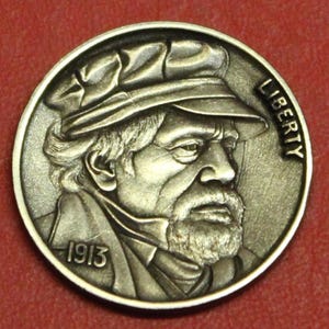 May include: Circular silver coin featuring a detailed profile of a man wearing a hat, with a beard and the year 1913. The word "LIBERTY" is inscribed along the right edge of the coin. The coin has a detailed, aged appearance.