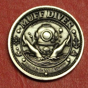 May include: A silver-colored coin with the words "MUFF DIVER" around the top edge. The coin features a diving helmet, hands reaching up from the water, and the text "Exploring the Unknown" and "Inspired by the Spirit of Adventure."