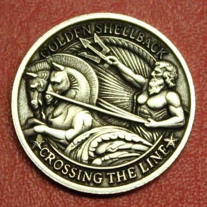 May include: A silver-colored coin with the words "GOLDEN SHELLBACK" and "CROSSING THE LINE" engraved around the edge. The coin features a detailed relief of a chariot pulled by two horses, with a figure holding a trident.