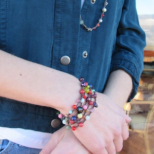 May include: A close-up of a person's arm wearing a multi-strand beaded bracelet with colorful beads and a dark heart charm. The bracelet is on a person's wrist wearing a blue denim jacket and a white shirt.