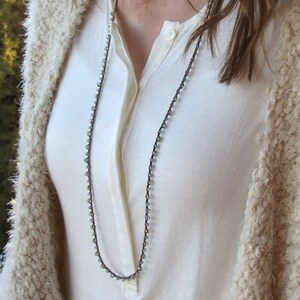 May include: A long, delicate necklace with small, light blue beads and a brown cord. The necklace is worn by a person with long brown hair and a white shirt.