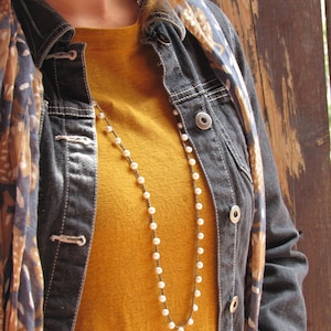 May include: A woman wearing a denim jacket with a floral scarf and a long pearl necklace over a mustard yellow shirt.