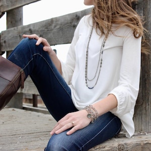May include: A woman wearing a white long-sleeved shirt, blue jeans, and brown boots. She is sitting on a wooden surface and has a layered necklace and bracelets on.
