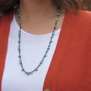 May include: A turquoise and brown beaded necklace with a simple design. The necklace is worn by a person wearing a white shirt and a rust-colored cardigan.