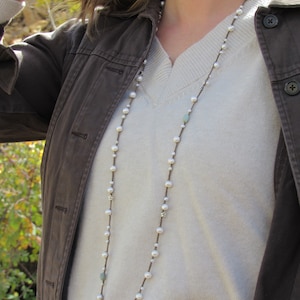 May include: A long white pearl necklace with silver accents and a light blue stone pendant. The necklace is worn by a person wearing a white sweater and a brown jacket.