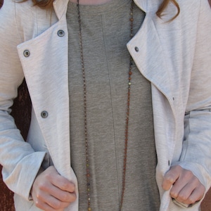 May include: A woman wearing a light gray jacket over a gray long-sleeved shirt with a long multi-colored beaded necklace.