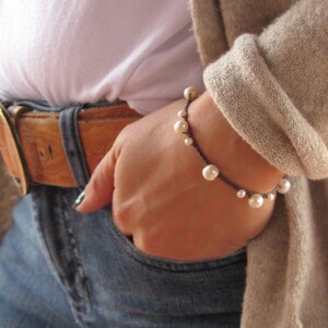 May include: A simple bracelet with white pearls and a brown cord. The bracelet is worn on a person's wrist.