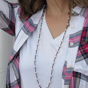 May include: A long brown and white pearl necklace with a simple design. The necklace is worn by a person wearing a plaid shirt and a white t-shirt.