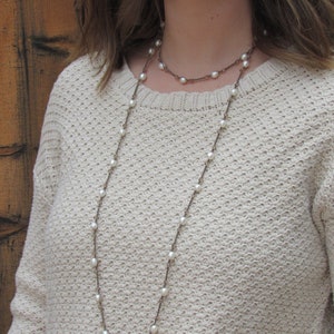May include: A long, layered necklace with white pearls and a brown cord. The necklace is worn by a person with long brown hair and a white sweater.
