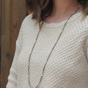 May include: A long, delicate necklace with small, white beads and a silver chain. The necklace is worn by a person wearing a cream-colored sweater.