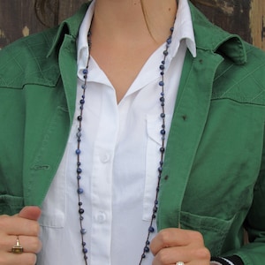 May include: A woman wearing a green jacket over a white button-down shirt with a long necklace of blue and brown beads.