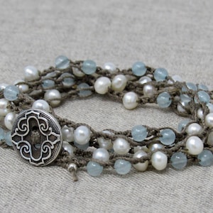 May include: A multi-strand bracelet featuring white pearls, light blue beads, and a silver button with an intricate design. The bracelet is made with brown crocheted cord.