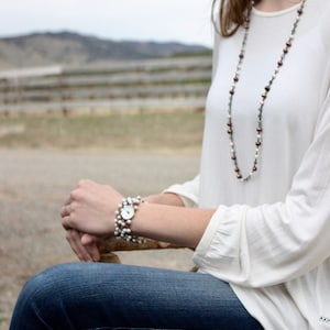 May include: A woman wearing a white long-sleeved shirt with a multi-colored beaded necklace and a pearl bracelet. She is sitting on a wooden surface with her legs crossed and wearing blue jeans.