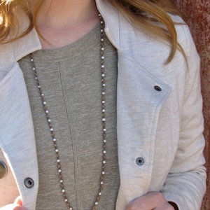 May include: A long brown and white beaded necklace worn over a gray t-shirt and a white jacket.