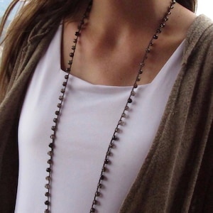 May include: A long brown beaded necklace with a simple design. The necklace is worn by a person with long brown hair and a white shirt.