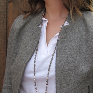 May include: A woman wearing a gray jacket and a white shirt with a long necklace made of gray and brown beads.