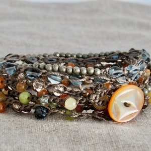 May include: A multi-strand bracelet with brown cord, beads, and a large, white button. The bracelet features a variety of beads in different colors and shapes, including clear, green, brown, and orange.