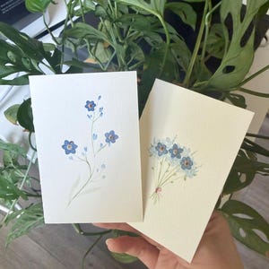 May include: Two watercolor greeting cards featuring blue forget-me-not flowers. One card displays a single stem with blossoms, the other a bouquet tied with a pink ribbon. The cards are on a white background.