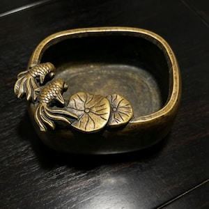 May include: A square-shaped bronze bowl with rounded corners featuring a decorative relief of two fish and two lily pads on the top edge. The metal has a dark, aged patina with golden highlights on the raised fish and lily pad details.