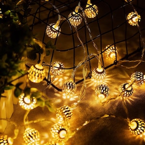 Puede incluir: Cadena de luces LED blanco cálido con un diseño decorativo de metal calado. Las luces están ensartadas en un cable fino y están dispuestas alrededor de una cesta de metal negro. El efecto general es un brillo suave y acogedor.