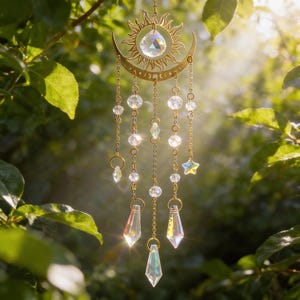 May include: A celestial-themed suncatcher with a gold-colored sun and crescent moon design. Clear crystal beads and prism pendants hang from the frame. The suncatcher is suspended among green leaves, catching the sunlight.
