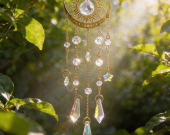 May include: A celestial-themed suncatcher with a gold-colored sun and crescent moon design. Clear crystal beads and prism pendants hang from the frame. The suncatcher is suspended among green leaves, catching the sunlight.