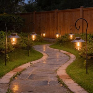 May include: A winding stone pathway illuminated by several hanging outdoor lanterns. The lanterns have a vintage design with a metal cage and a warm, inviting glow. The pathway is bordered by green grass and shrubbery, with a wooden fence in the background.
