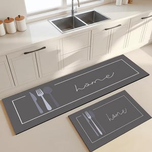 Op de afbeelding: Twee grijze keukenmatten met witte randen en het woord "home" in cursief. Eén mat is rechthoekig, de andere kleiner. Beide matten hebben silhouetten van keukengerei.