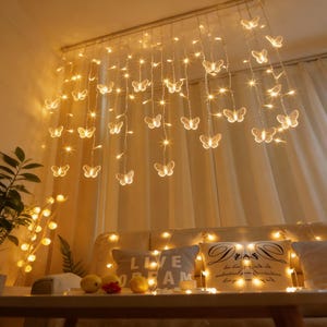 Op de afbeelding: Een decoratieve lichtslinger met transparante vlinders en warmwitte lampjes. De vlinders hangen aan het plafond en creëren een sprookjesachtige en betoverende sfeer. De lichten zijn over een bed gedrapeerd, waardoor een gezellige ruimte ontstaat.