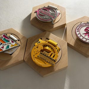 May include: Four medals displayed on wooden hexagonal bases. The medals are silver and gold, with colorful accents and text. One medal reads "SEMI MARATHON INTERNATIONAL de POINTE-A-PITRE". Another says "Les Foulées Ruban Rose".